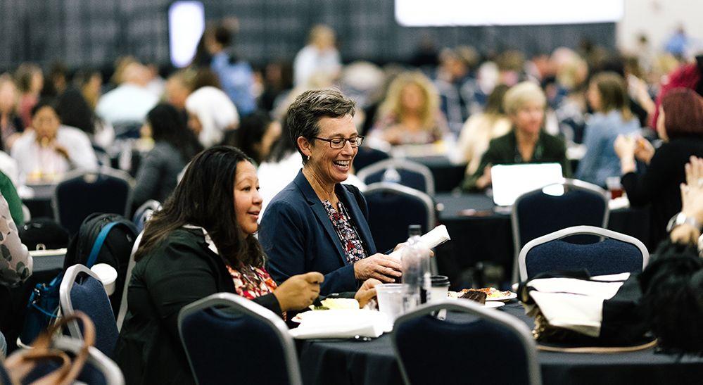large room of people at tables and two people smiling in response to something in the foreground