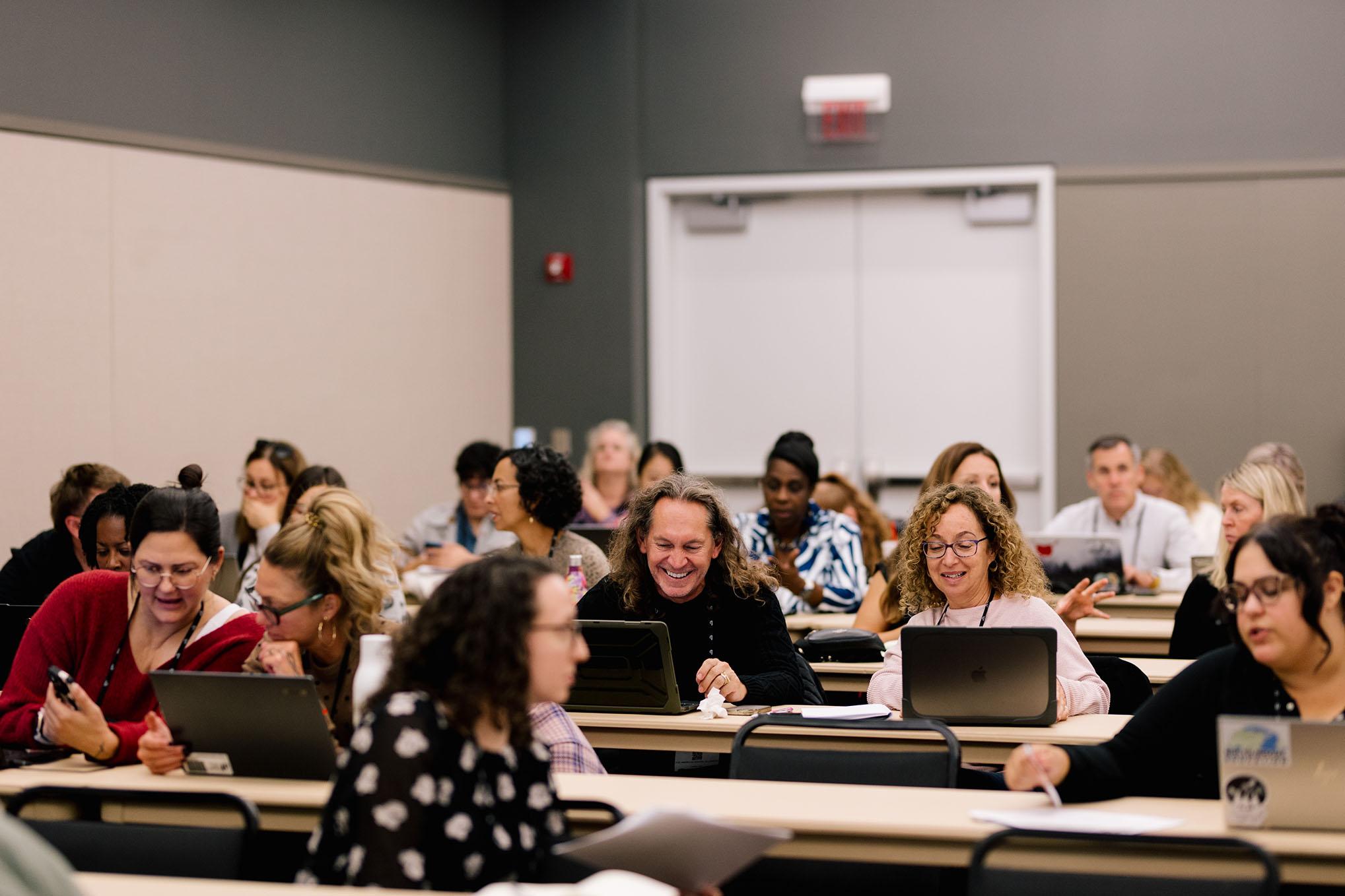 Conference attendees sit at tables in rows, talking and working together